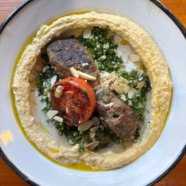 Hummus plate with two pieces of grilled meat, a grilled tomato, and green herbs, garnished with almonds.