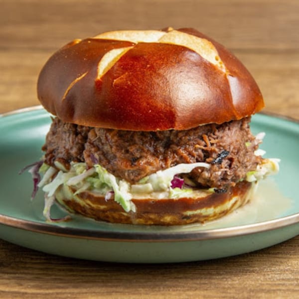 A Gili's Kitchen kosher pulled beef sandwich with coleslaw on a pretzel bun, served on a teal plate.