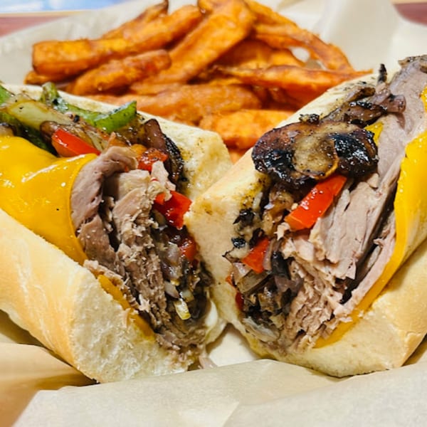 Two halves of a Gili's Kitchen kosher cheesesteak with sweet potato fries in the background.