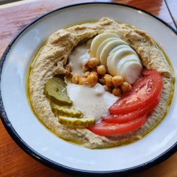 A bowl of Gili's Kitchen hummus topped with chickpeas, sliced egg, pickles, and tomatoes.