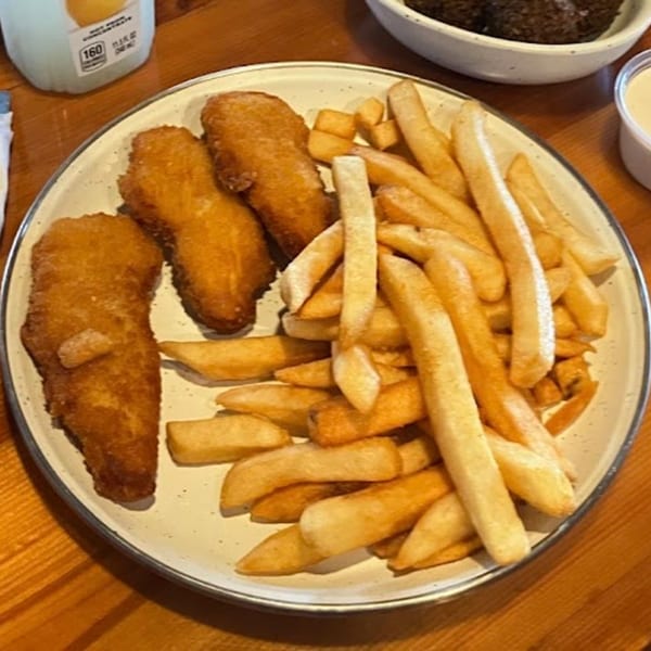 A plate of golden-brown fried fish and french fries, with a drink and sauce in the background.