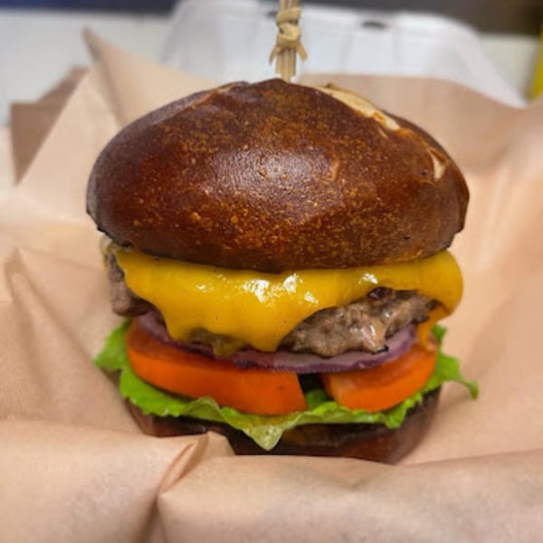 A Gili's Kitchen kosher burger with melted cheese, lettuce, tomato, and red onion on a pretzel bun.
