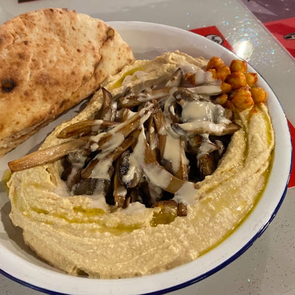 A bowl of Gili's Kitchen hummus topped with mushrooms, chickpeas, and a side of pita bread.